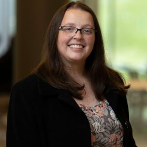 The image shows a professional portrait of a woman with long, dark brown hair and glasses, smiling warmly. She is wearing a floral-patterned blouse under a black jacket, and the setting is softly lit, with an out-of-focus background featuring natural light and hints of greenery. The photograph conveys a friendly and approachable demeanour, suitable for professional or academic contexts.